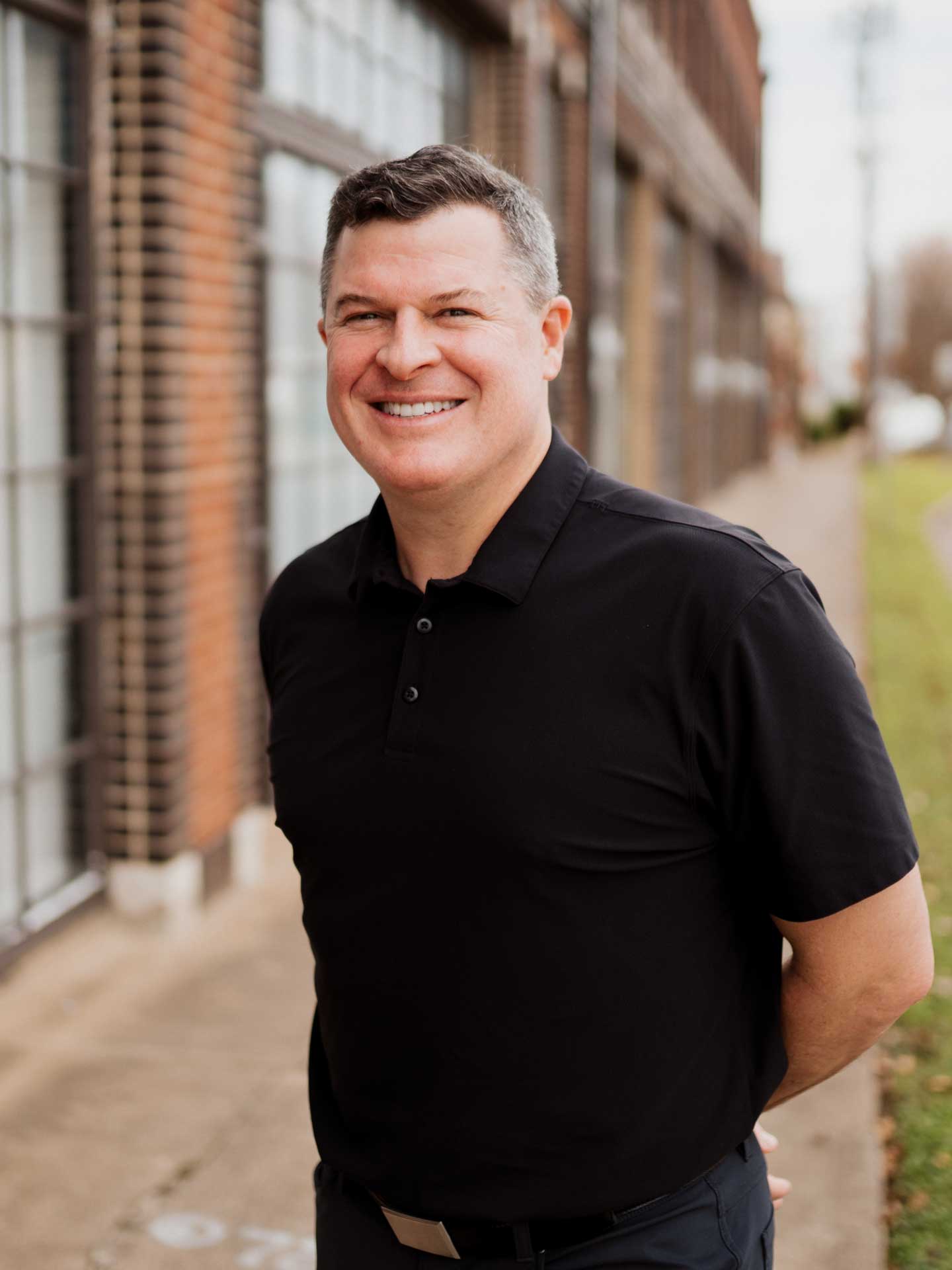 Patrick Fontana smiling with hands folded behind back in front of a brick wall wearing a black polo shirt